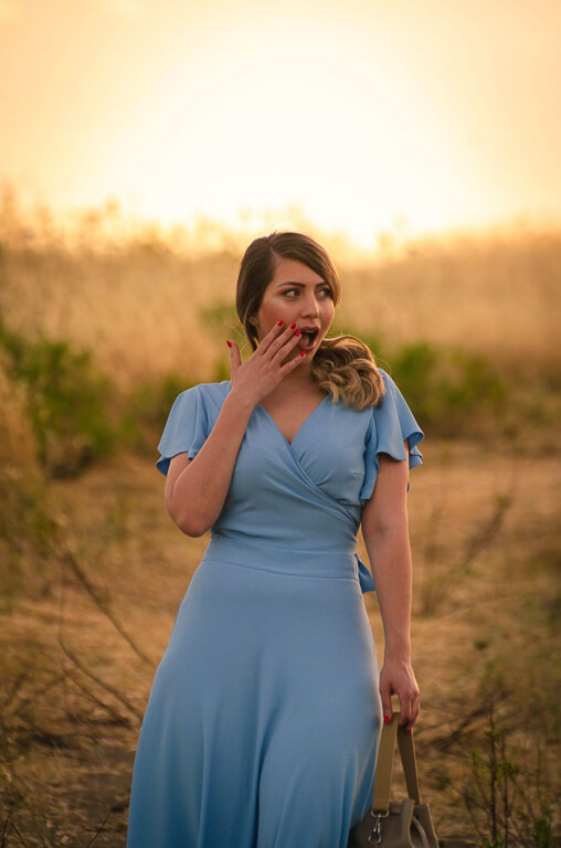 a woman looking down holding a beige bag in a blue dress and blue shoes in a field