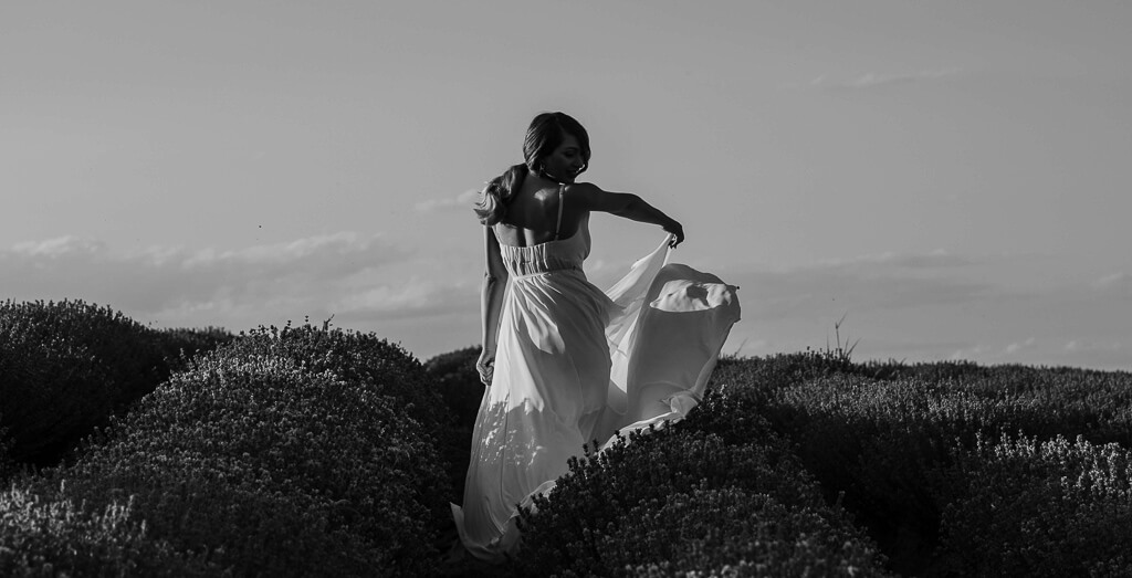 a woman in a yellow long dress is looking at the plain in a field of purple flowers and a blue sky