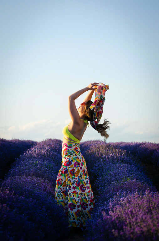 a woman in a yellow dress with her arms raised looks up as the sun embraces her. Στην A Collection Boutique.