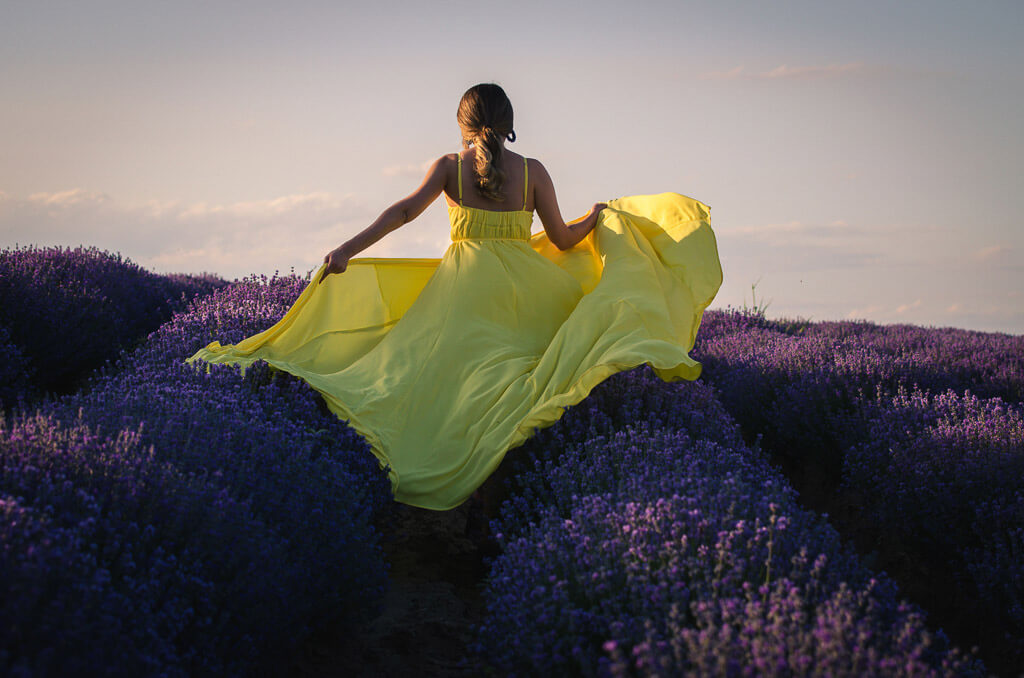 a woman runs holding a yellow dress outside among purple flowers and a blue sky