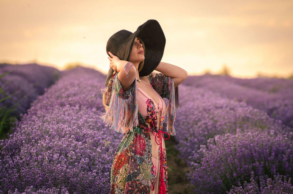 a woman holding her summer hat and wearing a colorful dress outdoors among purple flowers