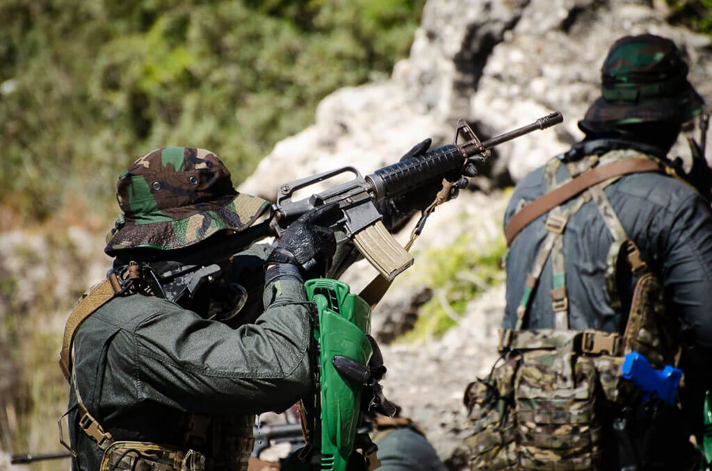 a soldier in uniform is holding an automatic weapon and pointing it. He's wearing a hat and a mask