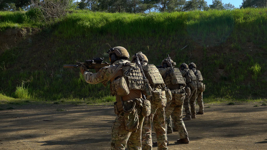 six soldiers in line with uniforms, helmets, and automatic weapons pointing while on a mountain