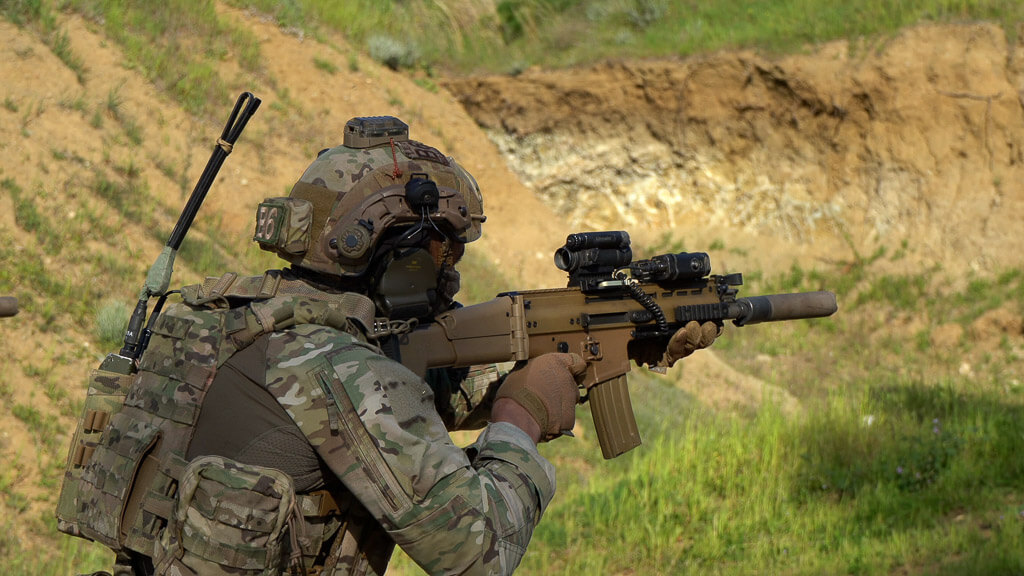 soldier in uniform, wearing a helmet, holding an automatic weapon, and aiming while on a mountain