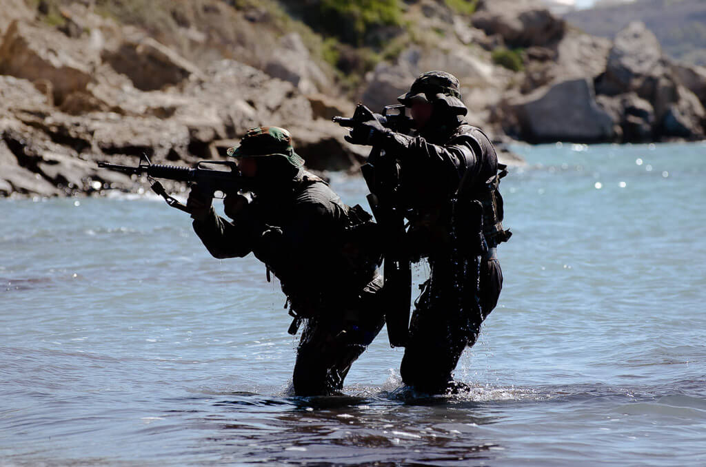 Two soldiers in uniform, hats, holding automatic weapons, and pointing while in a river.