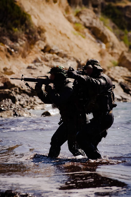 2 soldiers in uniform holding automatic weapons and aiming while in a river