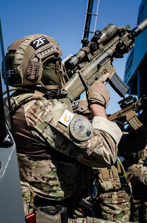 Α soldier in uniform holding an automatic weapon and aiming high. He's wearing a helmet and a mask.