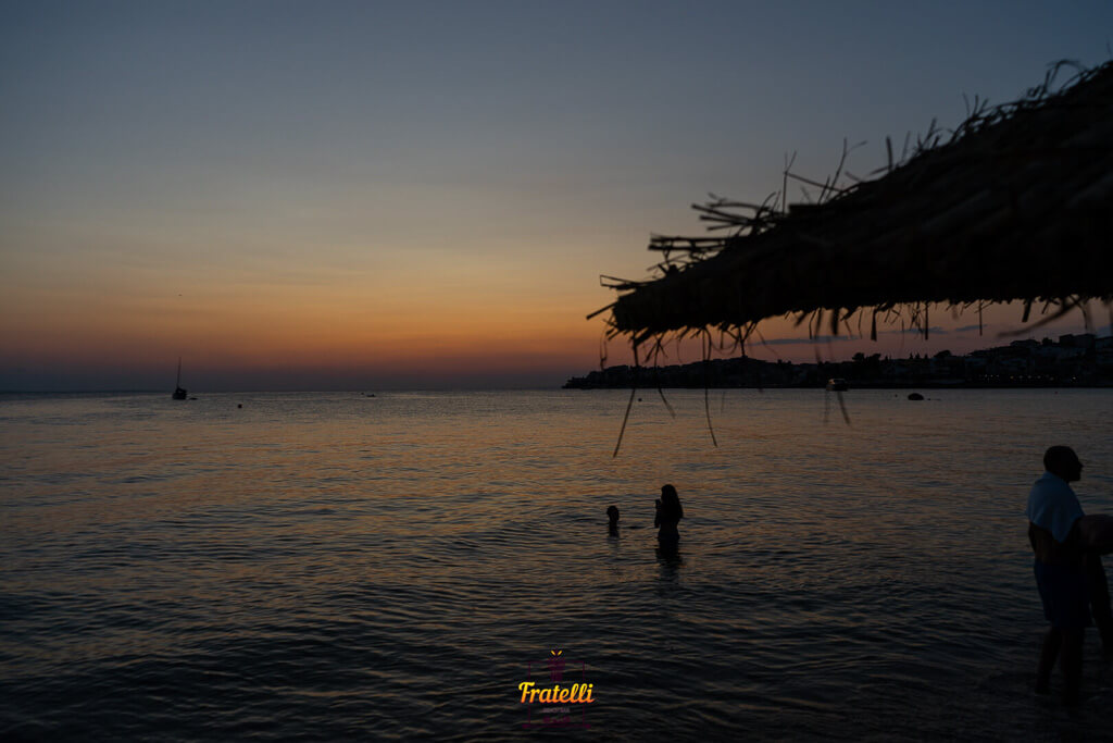 sunset and sea with two people in it cooling off. On the side is a straw umbrella