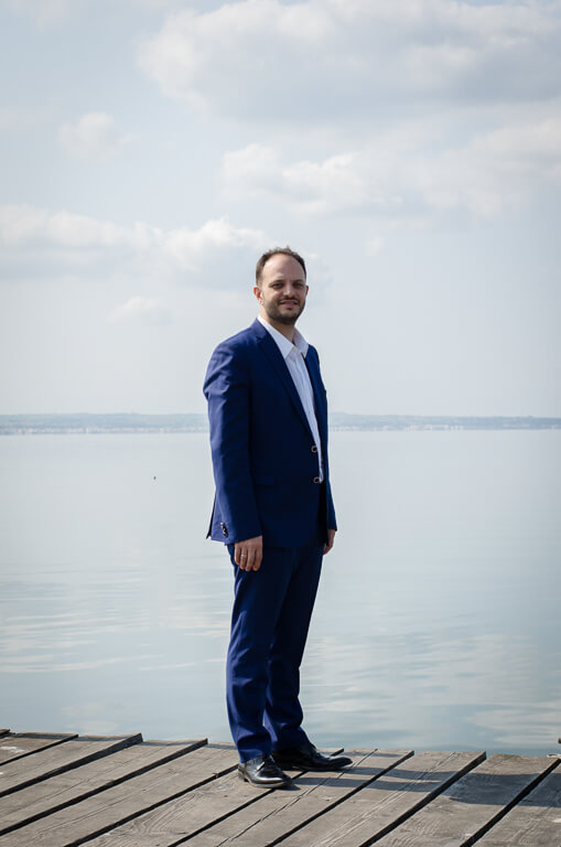 a man in a blue suit and white shirt on a wooden pier with the sea behind him.