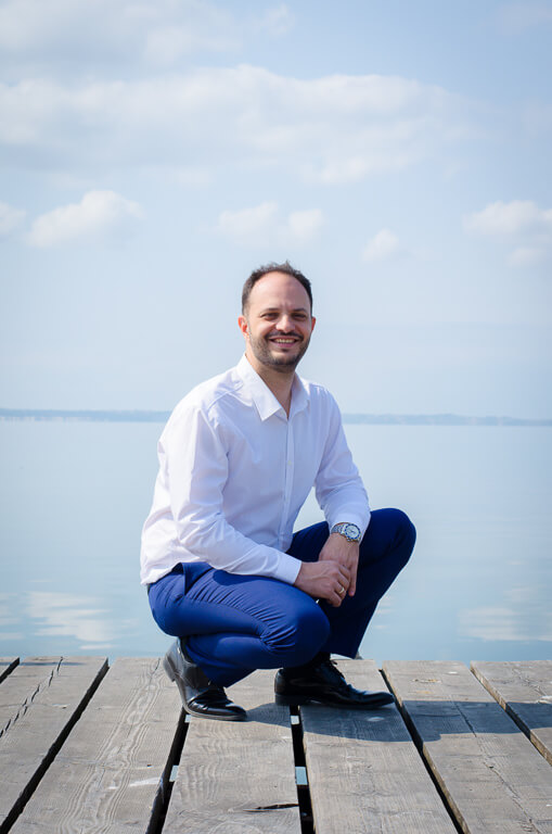 man smiling in blue trousers and a white shirt sitting on a wooden pier with the sea behind him.