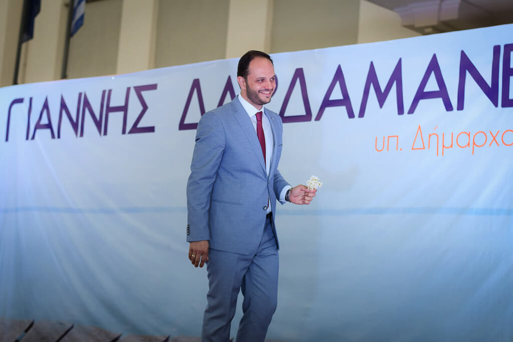 Mayor Dardamanelis in a grey suit, holding a white flower and walks on stage smiling.