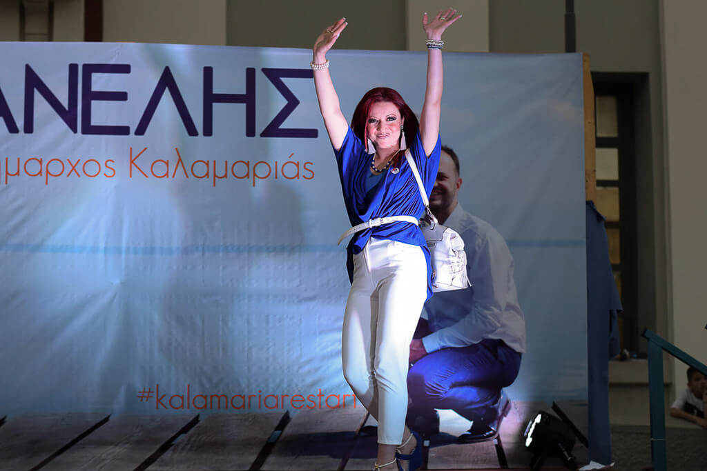 red-haired woman on stage smiles and waves. She's wearing white pants, a blue shirt, and a white bag