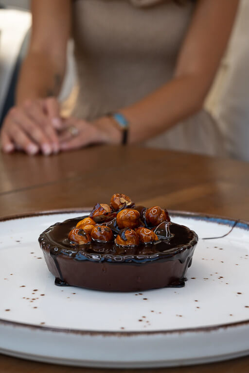 a white plate of chocolate dessert with hazelnuts and chocolate syrup on a wooden table.