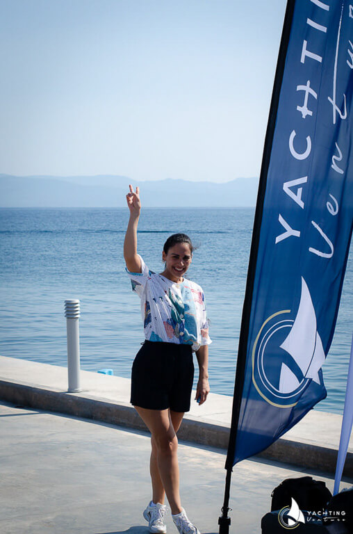 a girl smiles raise her right hand and make the peace sign, and next to her is a blue flag