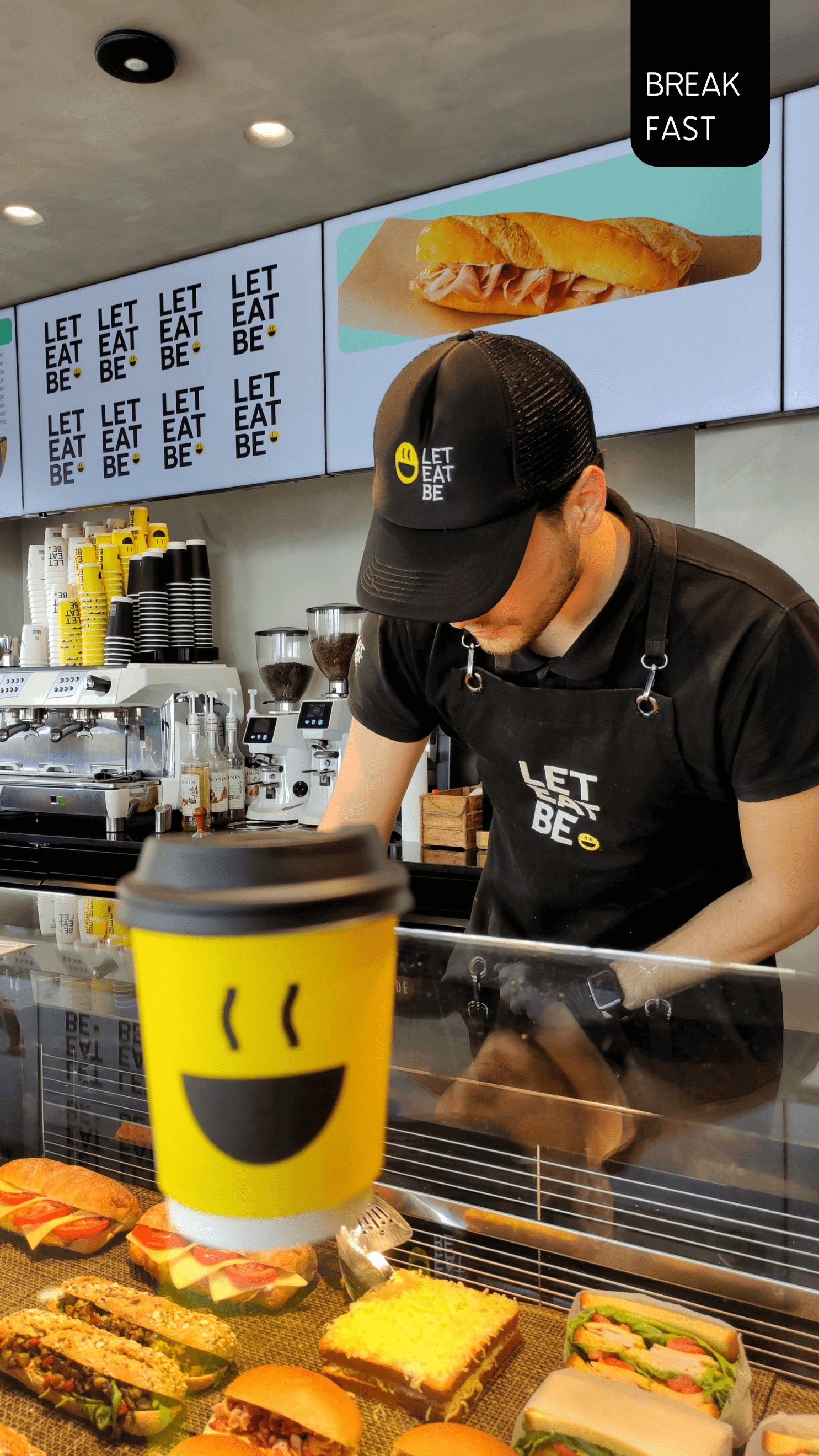 pass a restaurant that has food and croissants. upstairs in the window is a coffee cup in white, yellow and black and an employee in a black uniform is putting food in it. In the back there are displays of food and the name of the restaurant "Let Eat Be''.